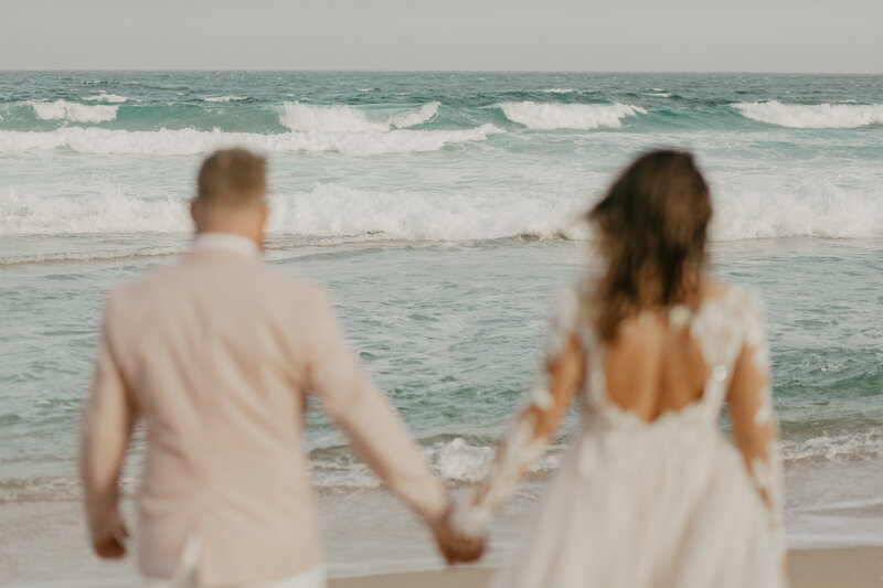 Wedding Photographer, a bride and groom in dress and beige suit, hold hands walking toward the ocean