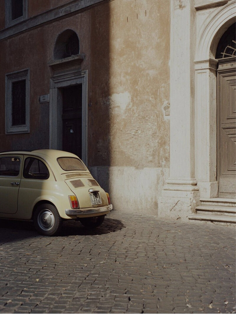 Quaint European alleyway with a vintage car exiting past stone walls.