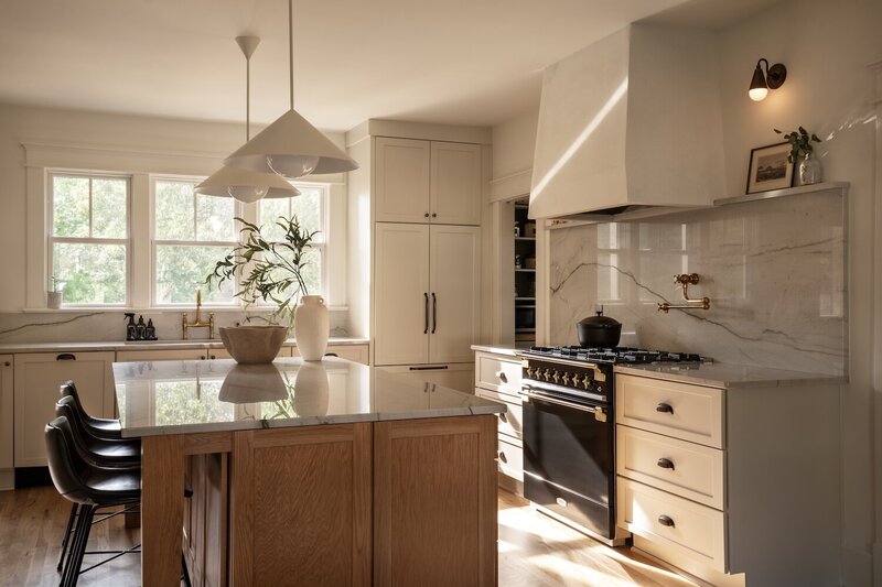 Gourmet chef kitchen with custom black and gold oven, white modern range hood, large marble backsplash, massive island with counter seating, and natural light from backyard windows.