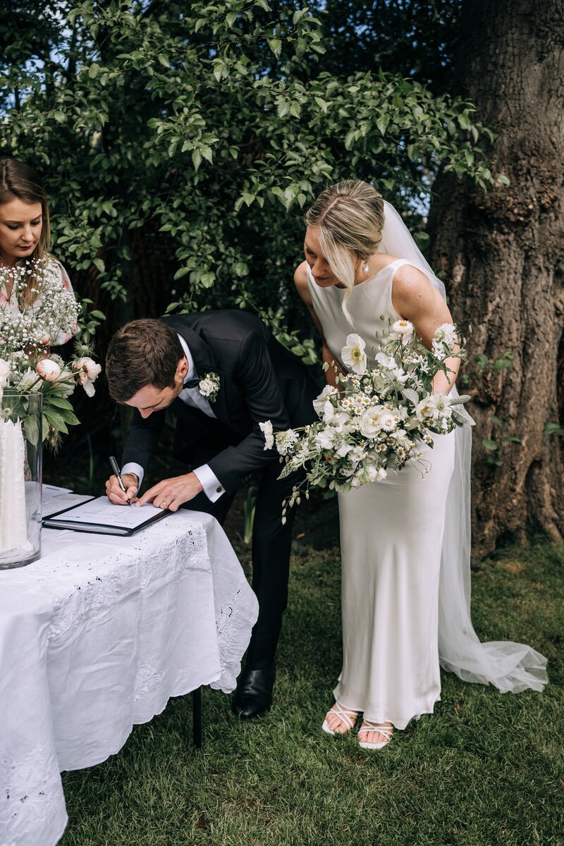 a groom signs marriage licence while his wife holds her white and yellow bouquet from mrs bottomleys flowers and watches