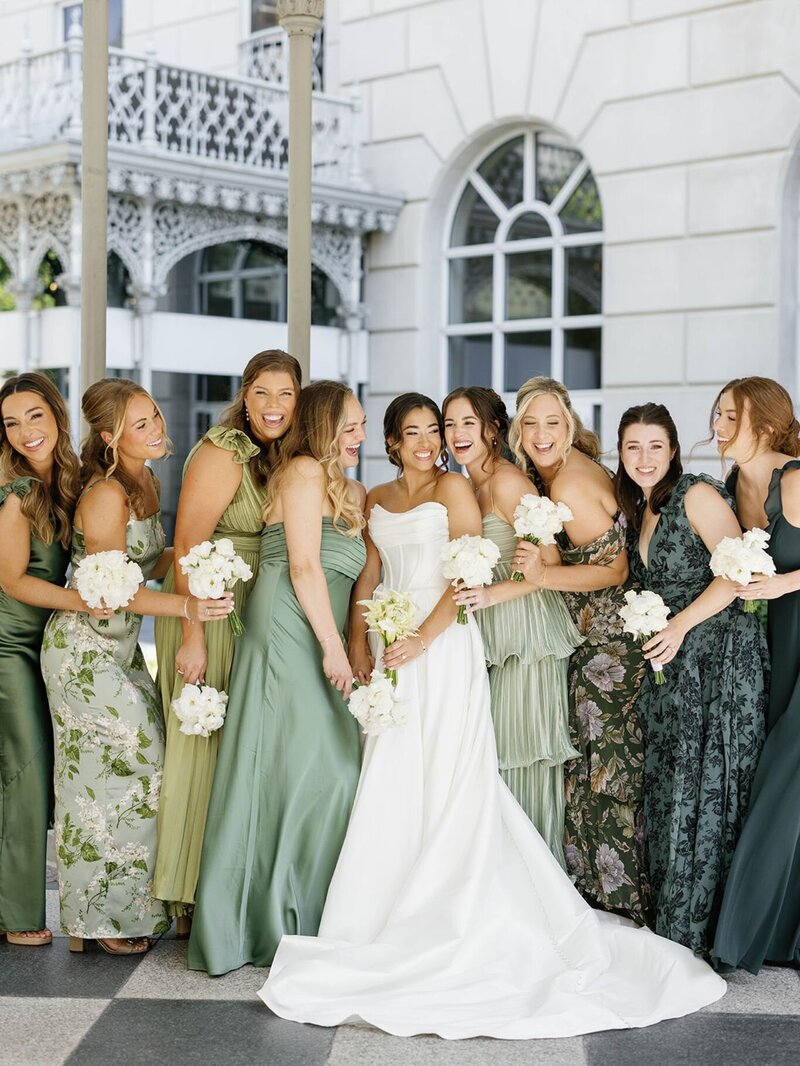 Bride surrounded by her bridesmaids wearing green and floral dresses, laughing together before the wedding ceremony.