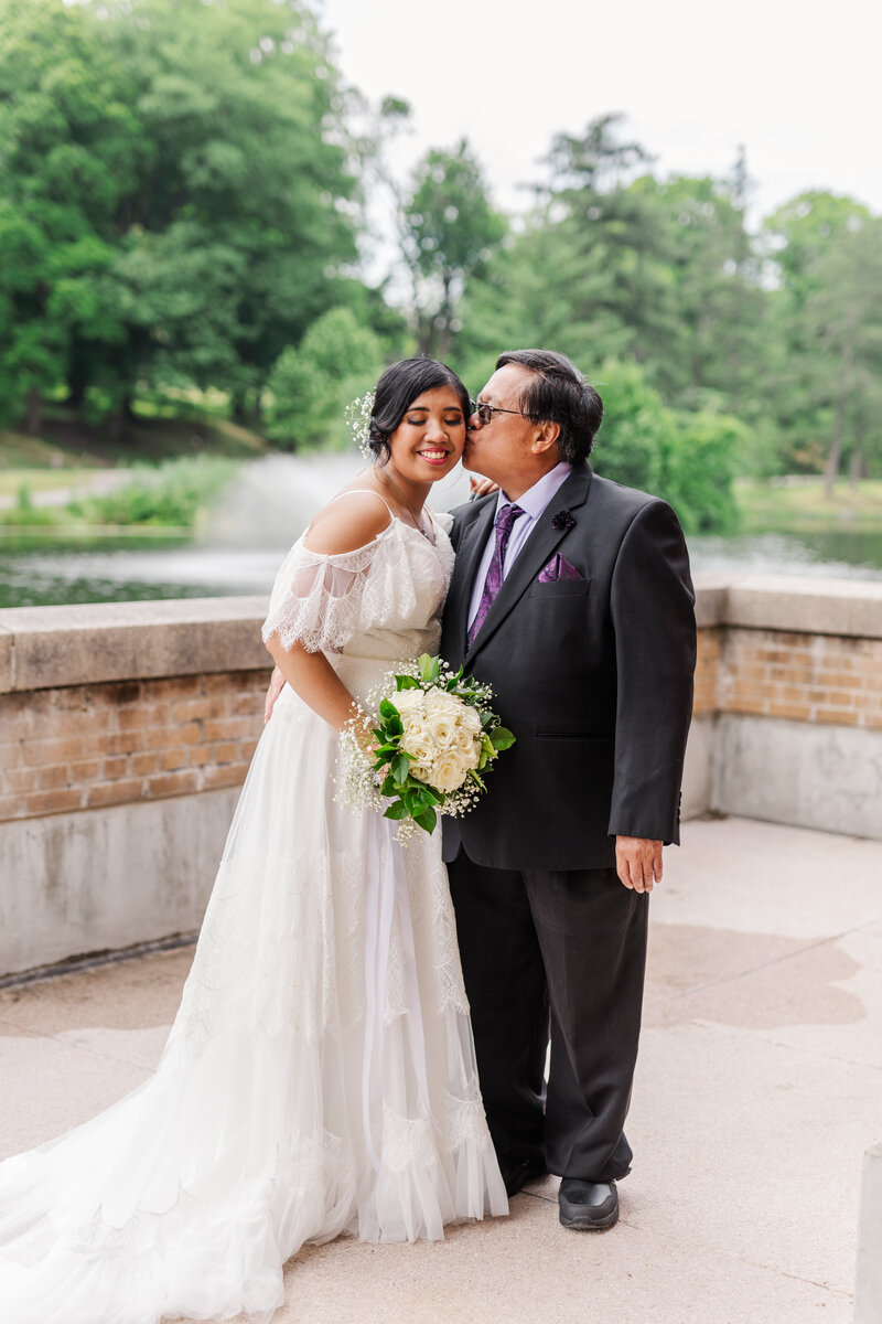father kissing bride on cheek with bouquet in park with water fountain during wedding photo session in albany ny 