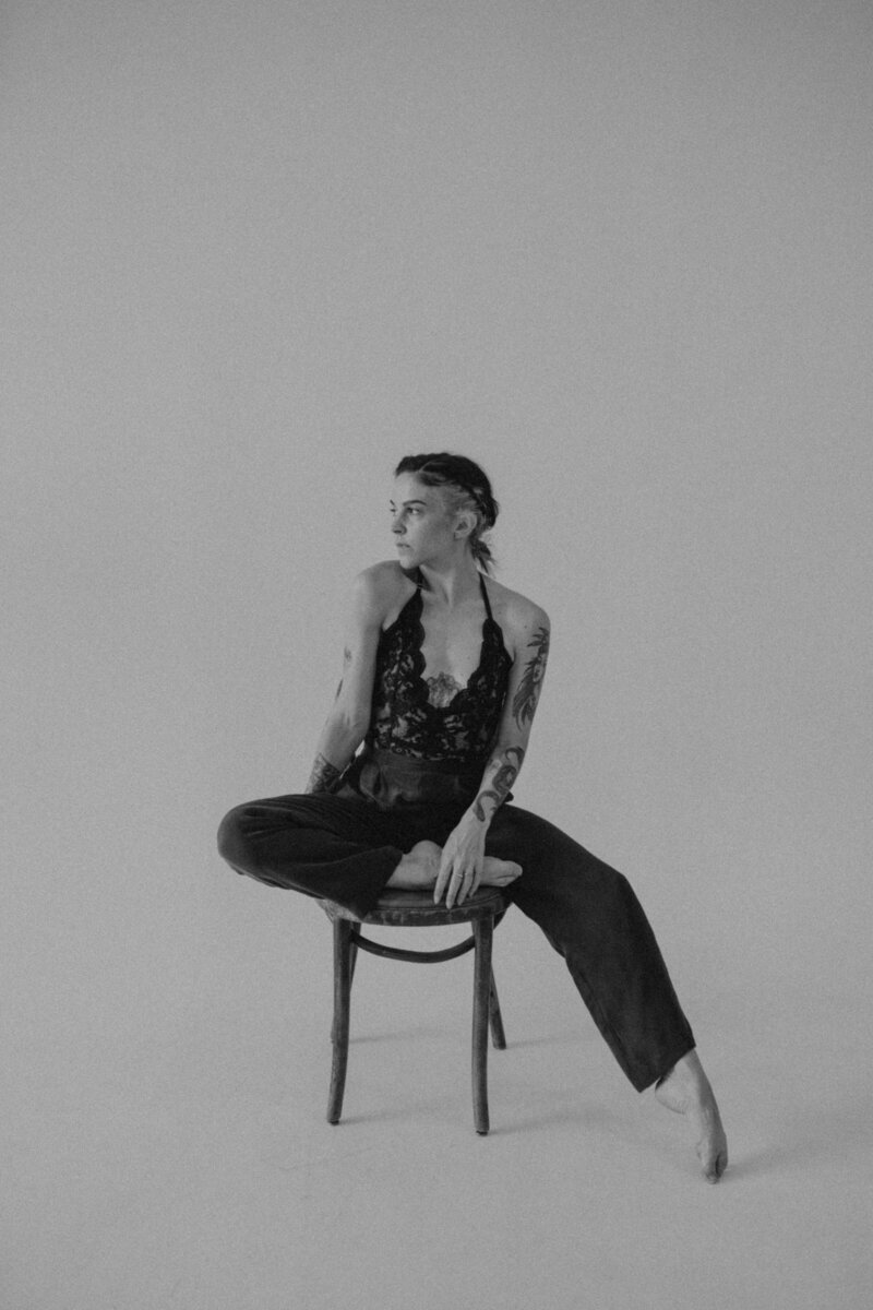 Black and white portrait of the photographer sitting cross-legged on a chair, barefoot in wide-leg pants and a lace halter top, in a minimalist studio.