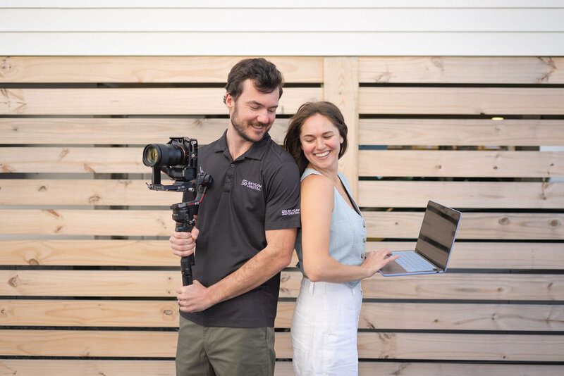 couple stand back to back as man holds camera and woman holds laptop