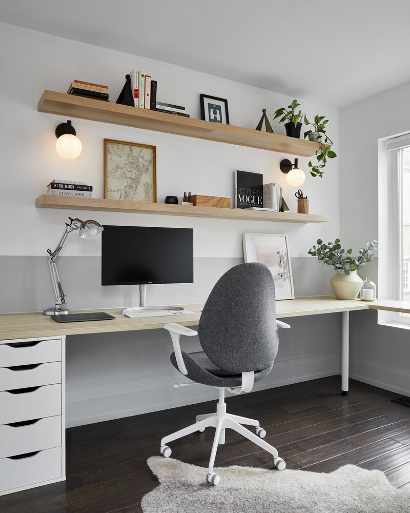 Before and after transformation of a simple bedroom layout into a vibrant, modern office space featuring a long wood desk with white drawers, grey and white rolling desk chair, floating wood shelves containing various books, art pieces, and potted plants. The white walls and throw rug contrast with the dark brown hardwood flooring.