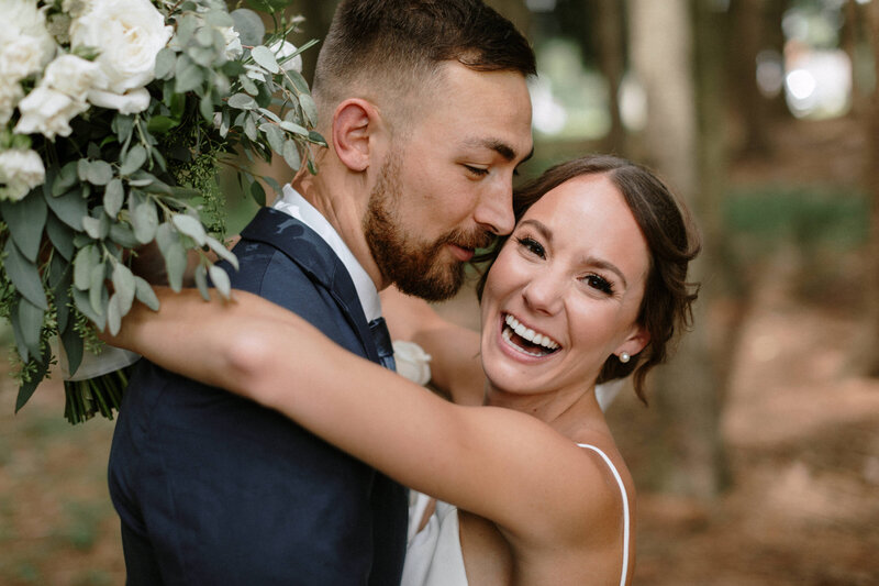 A couple embraces in a forest, surrounded by greenery, with one partner holding a lush bouquet of white roses and eucalyptus leaves.
