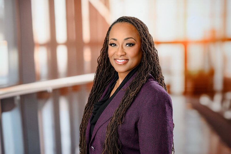 A woman in a purple suit jacket and long dark hair with a office style studio background