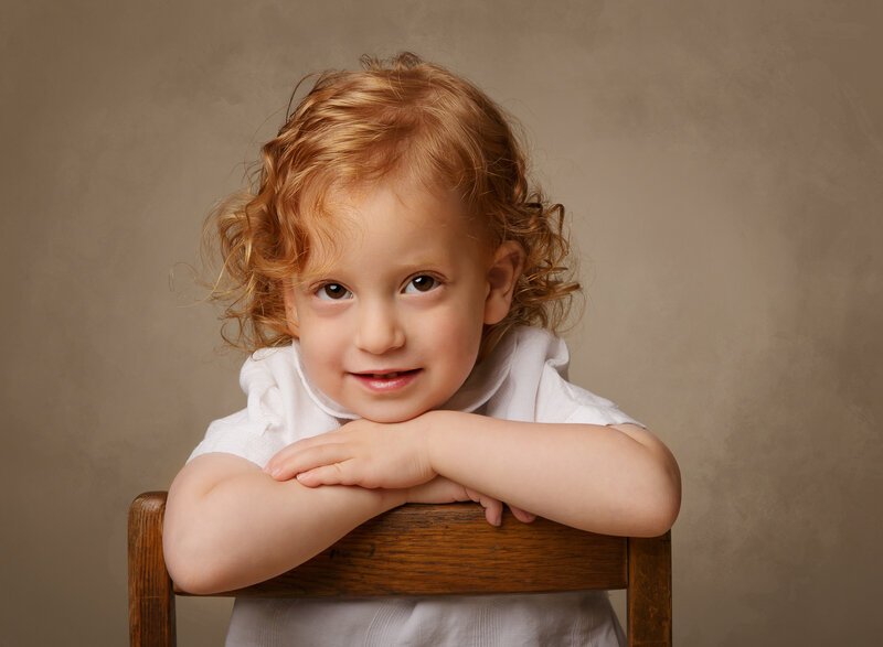 Brooklyn family studio photoshoot. Little boy with curly red hair is sitting backwards on a chair with his hands folded on the back and his chin resting atop. He is smiling at the camera. Captured by best Brooklyn, NY family photographer Chaya Bornstein.