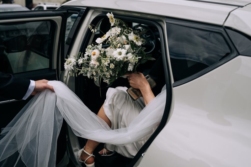 a hand holds a brides veil as she gets into a white car with her green white and yellow wild bouquet