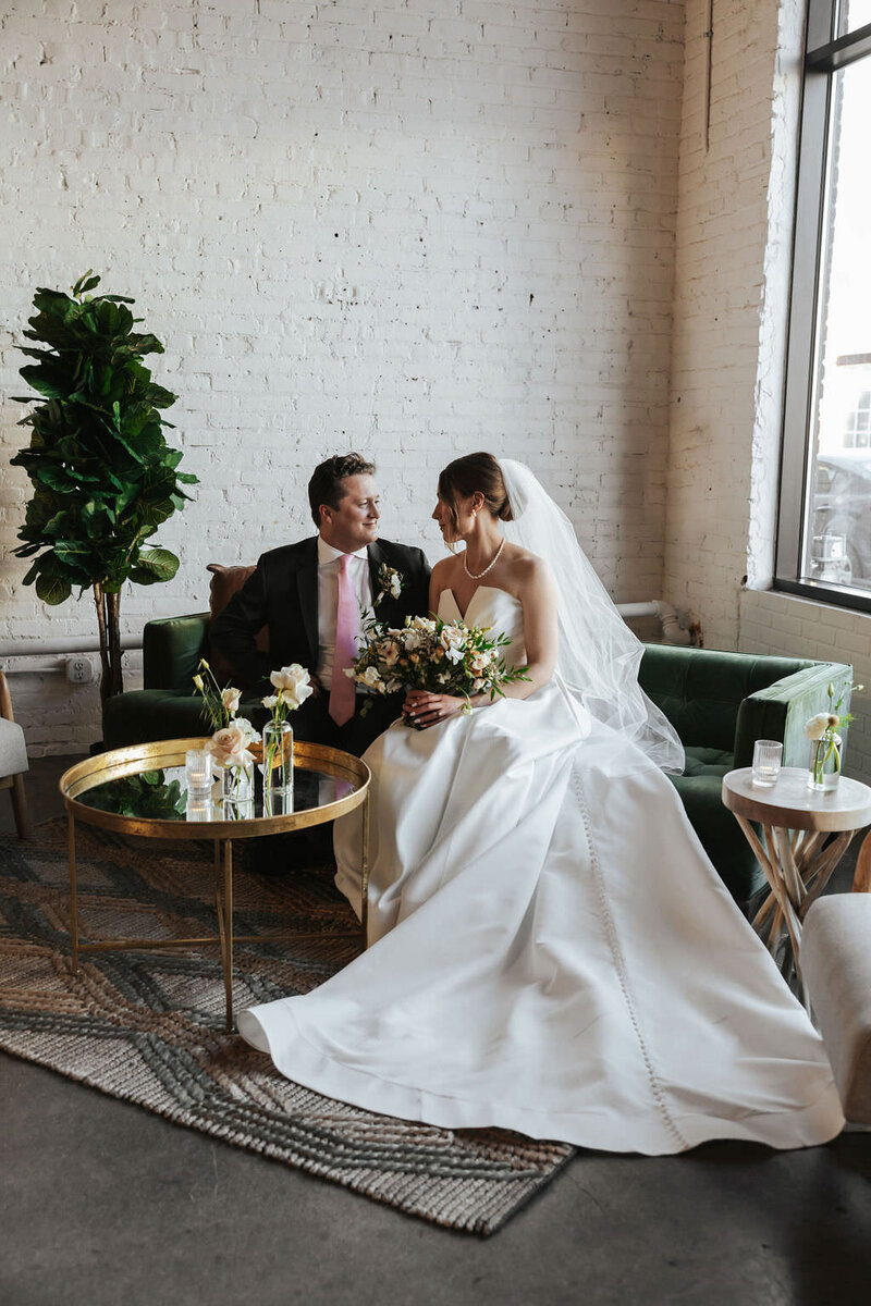 A newlywed couple sitting on a couch while one holds a bouquet of flowers