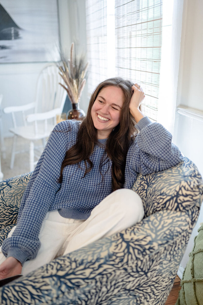 Annie wears a blue sweater sitting on an arm chair with all of the coastal vibes
