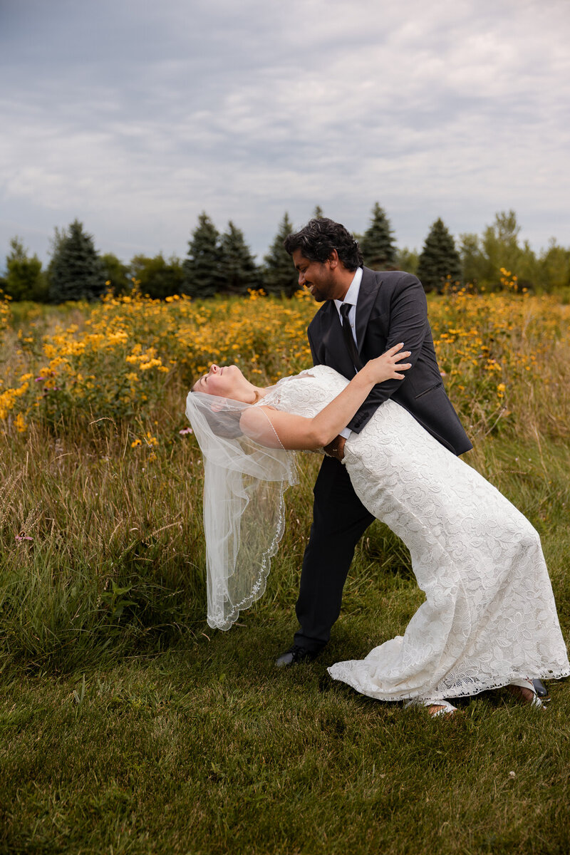 Bride and groom posing for a photo in a field of wild flowers and tall grasses. The field is surrounded by pines. The groom is dipping the bride back and smiling at her.