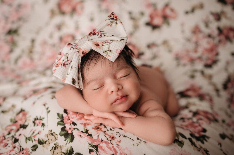 Newborn girl laying on tummy with legs and arms tucked under her chin and laying on floral backdrop and wearing a big floral bow