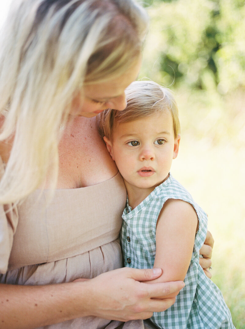 2 year old blonde boy in green gingham snuggles into his kneeling, blonde mother's chest as she comforts him.