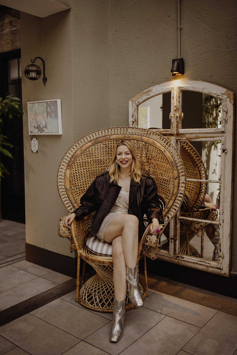 Lauren sits in a large wicker throne, she's wearing a black leather jacket and nude dress.