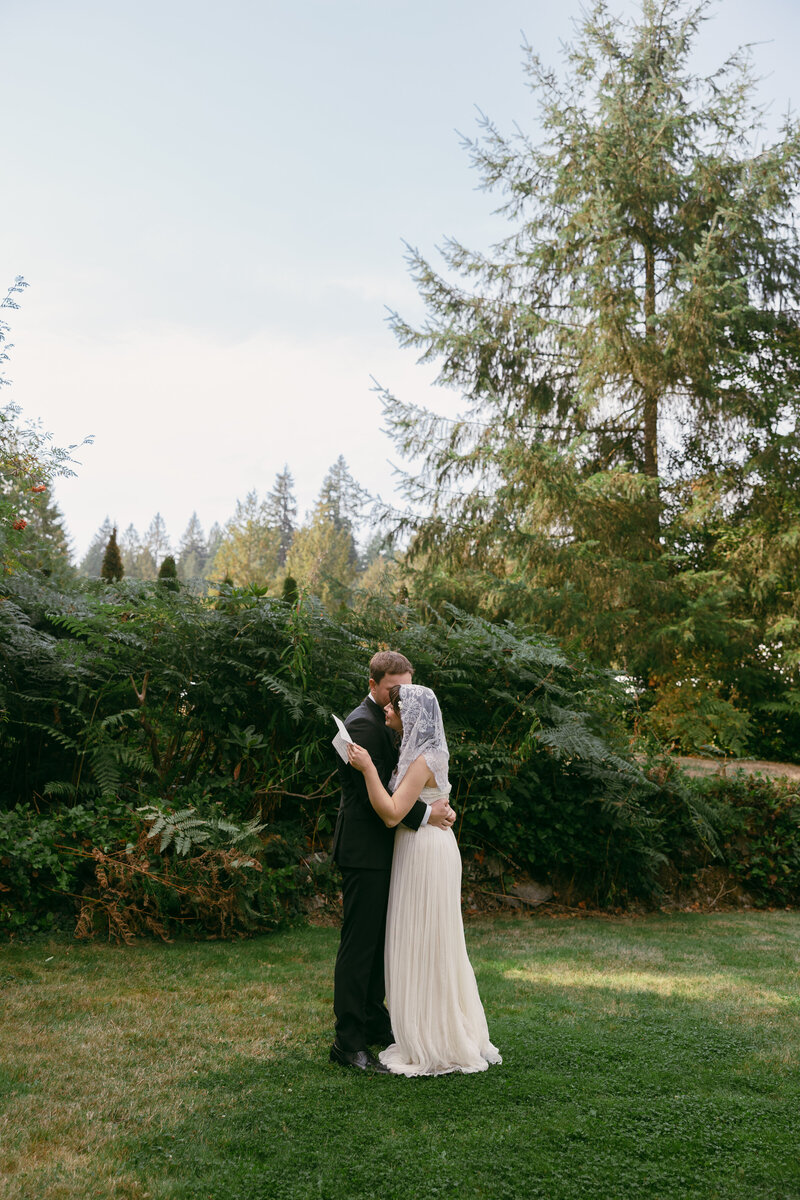 A man in a black suit holds a woman in a white dress with a lace veil over her head surrounded by greenery