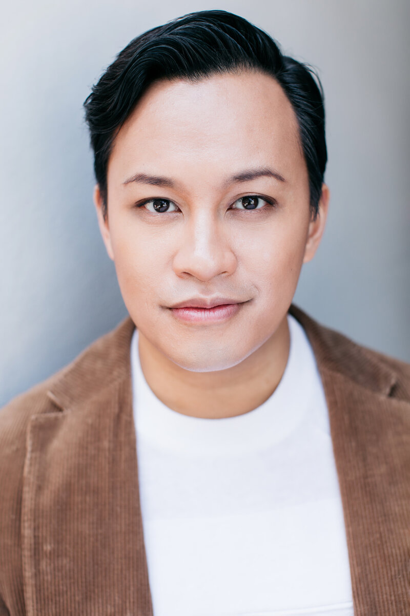 Casting headshot van man met kort zwart haar, wit shirt en bruine jas voor grijze muur.