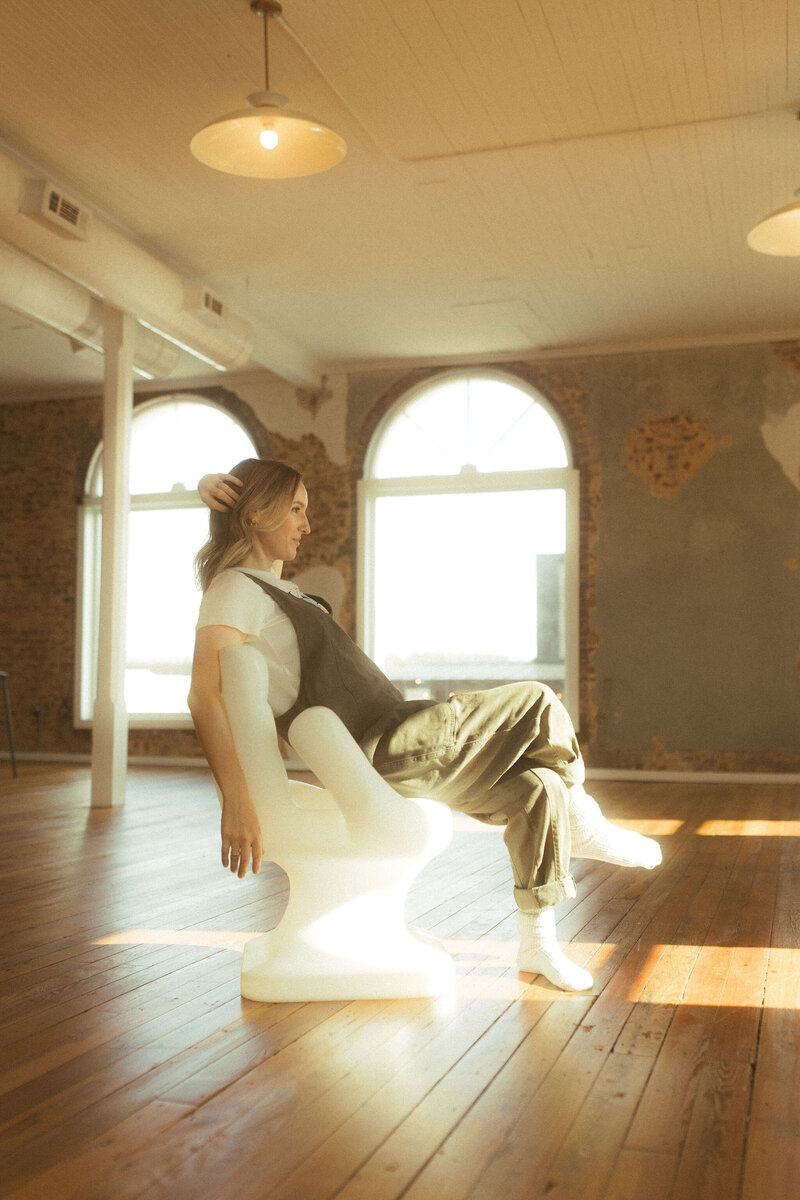 A woman in green overalls sits in a hand shaped chair and looks out the window. 