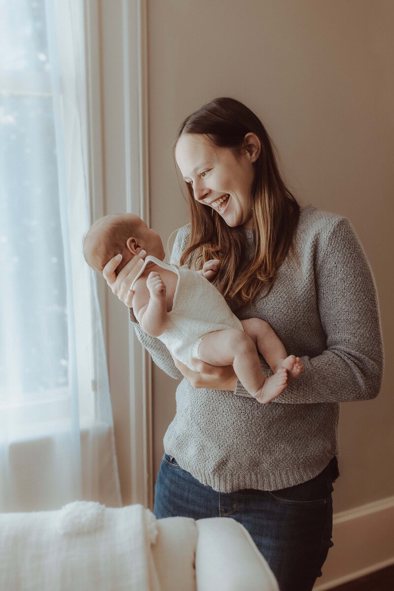 A woman in a gray sweater holds a smiling baby in a white onesie by a window with sheer curtains, capturing the warmth and tenderness of this moment. As if orchestrated by a Macon newborn photographer, the scene is bathed in soft natural light, accentuating their joyful bond.
