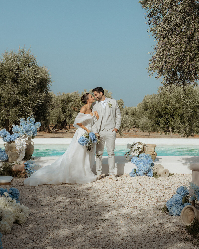 Bride and groom share a joyful, intimate moment in front of a serene pool, surrounded by lush olive trees and Mediterranean-inspired blue and white floral arrangements at Masseria Moroseta in Puglia, Italy.