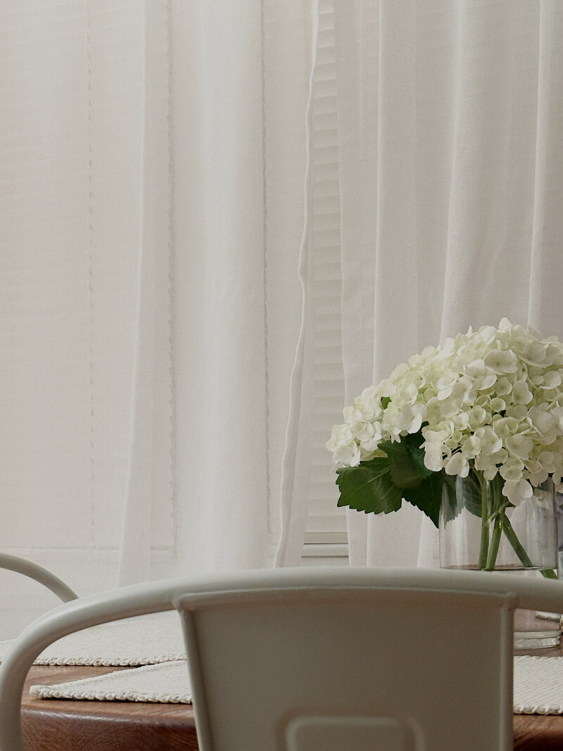 A serene dining area with a white metal chair, a wooden table, and a vase of white hydrangeas. Soft natural light filters through sheer curtains.
