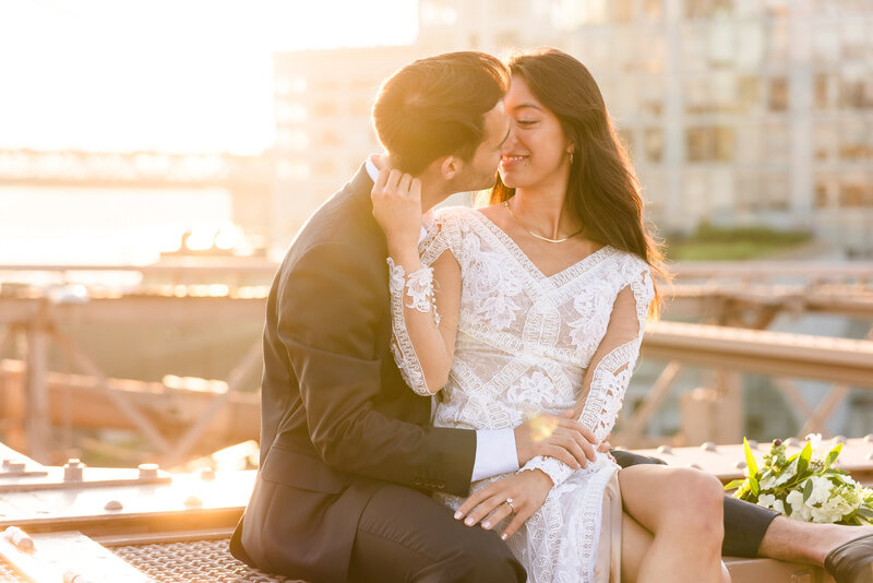 Bride and groom share a kiss at golden hour on the Brooklyn Bridge, her lace dress glowing in the light, captured by NYC wedding photographer Heritage House.
