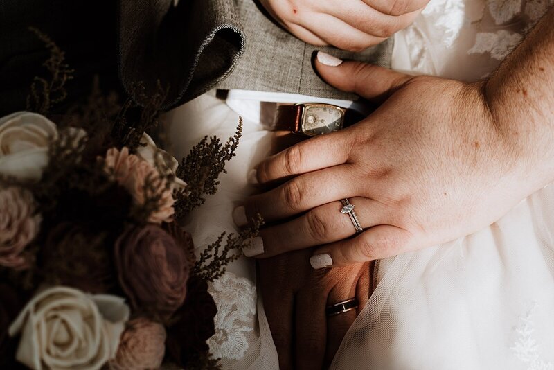 bride's hand lay on top of groom's hand showing her wedding ring and him showing his watch