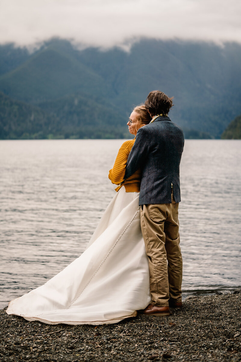 A couple in their wedding attire and cozy sweaters stands on the beach of Lake Crescent embracing after their Olympic National Park elopement