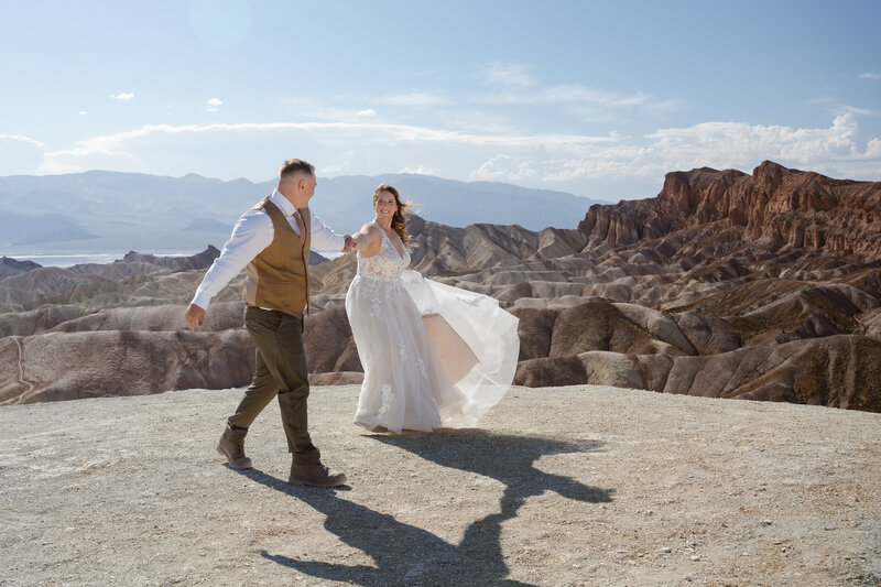 A man and woman hold hands smiling at each other in front of the rock formation called Zabriskie Point 