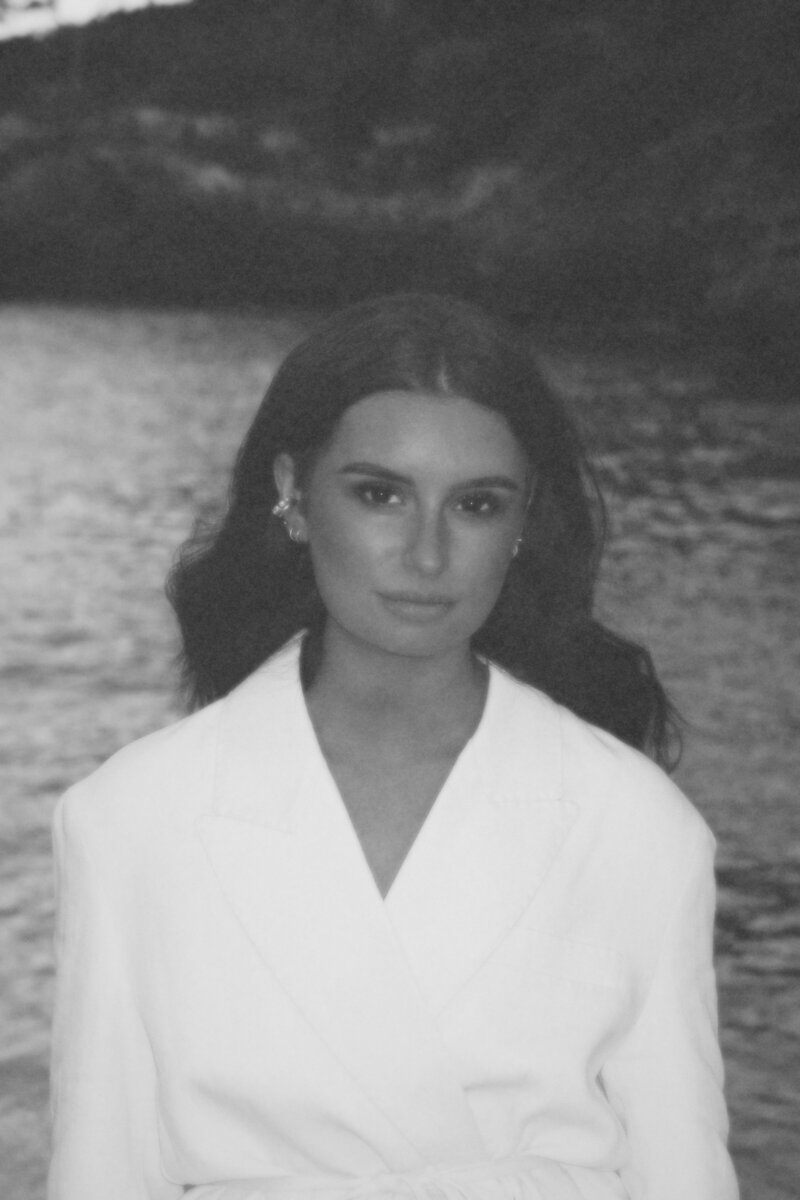 Black and white portrait of Studio Petita founder Chloe Harrison standing near calm water, wearing a structured white blazer, with soft waves in the background. The image conveys a sense of calm, elegance, and intentionality.