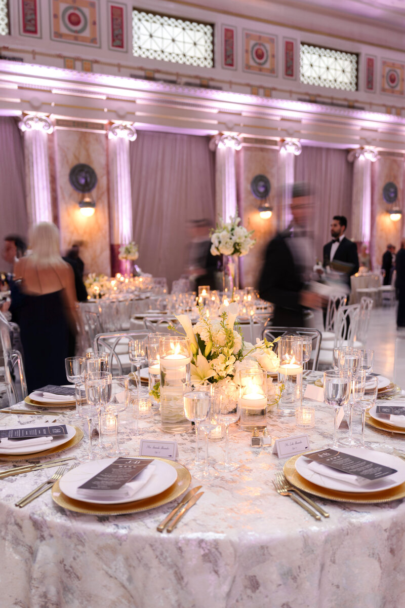 A beautifully set wedding reception table with elegant floral centerpieces, candles, and neatly arranged plates and menus. The background features ornate architectural details and softly lit columns, with guests mingling in formal attire.