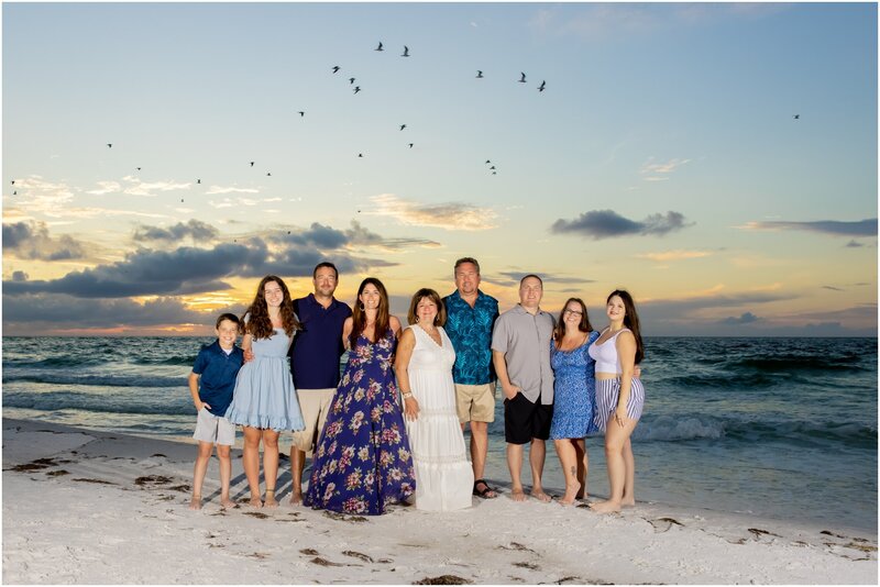 A family portrait of nine on the beach with the sun setting and birds flying behind them