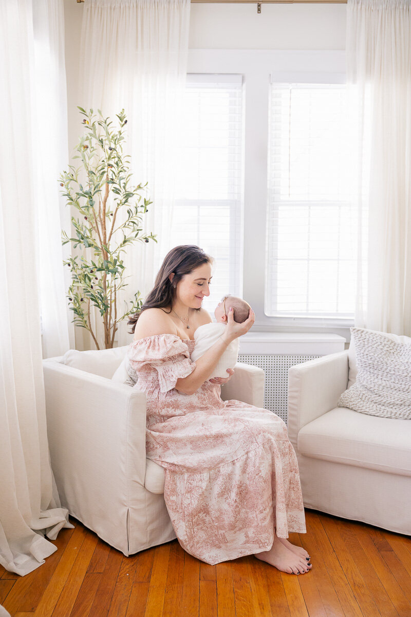 A mother in a soft pink dress lovingly holding her newborn in a sunlit, cozy living room with neutral decor and natural light streaming through tall windows.