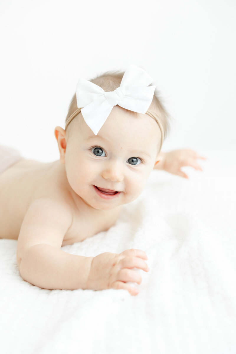 Smiling 6 month old baby girl wears white boy headband and lays on her belly during baby milestone portrait session