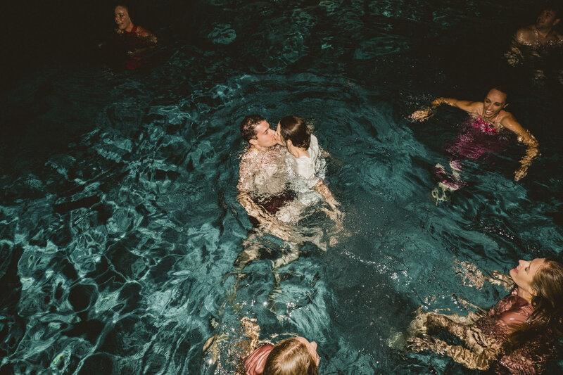 A bride and groom having a nigh-time swim and kissing in a blue pool, surrounded by wedding guests.