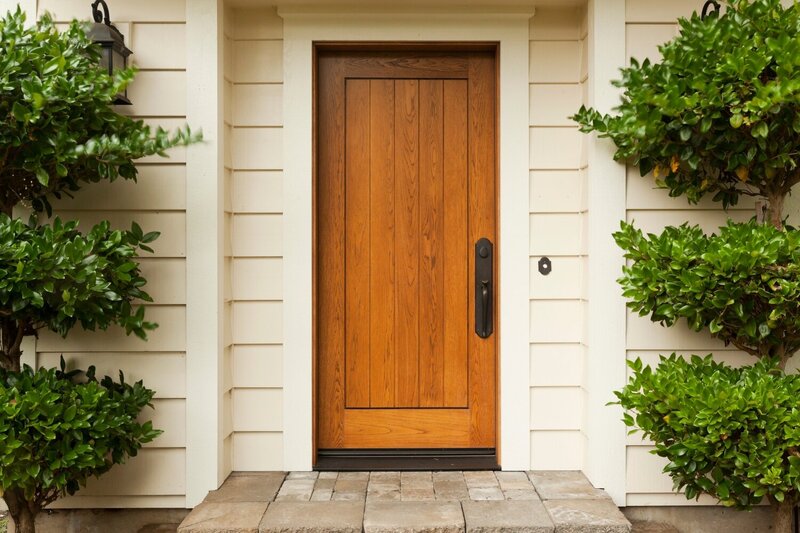 A warm oak paneled front door surrounded by creamy white lap trim with two topiaries on either side