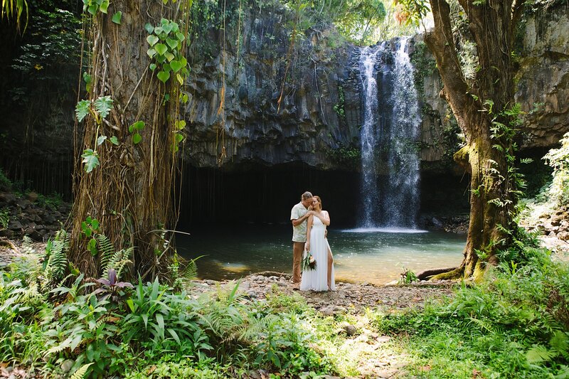 A bride and groom stand together in front of a large waterfall in Hawaii during their elopement session