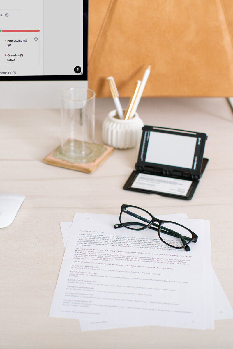 Printed wedding shot list and reading glasses on a clean white desk with a monitor and keyboard
