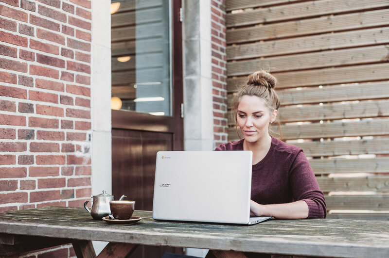 A white woman sits at a picnic table. There are wooden blinds behind her and a brick building with a dark wooden door next to her.  She's got her brown hair in a bun and has a Maroon shirt on. She's typing on her computer with a brown coffee mug and a silver pitcher of cream next to her