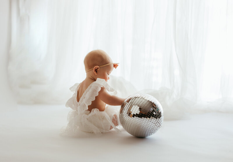 An eight month old baby girl dressed in a white lace dress sits on an all white backdrop with sheer curtains as she plays with a disco ball sitting to her side during her Denver milestone session