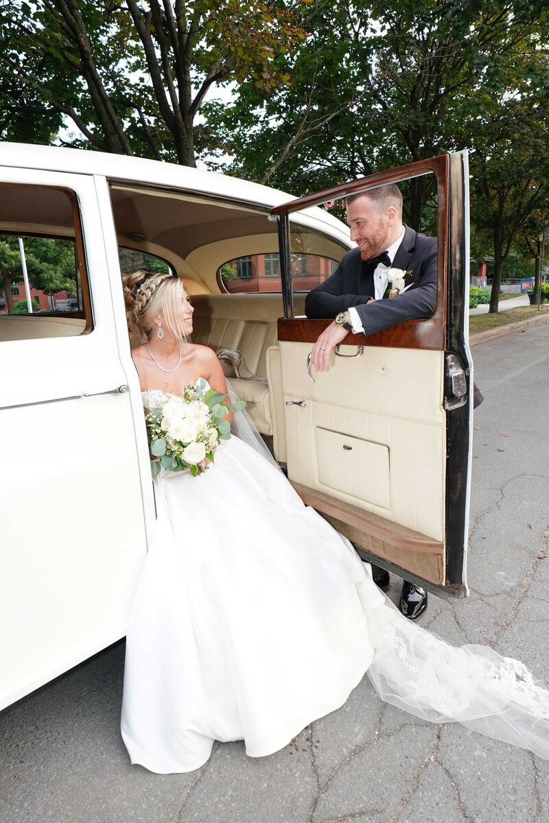 Une mariée est assise avec élégance dans une voiture, la porte ouverte, tandis que son époux s'appuie sur la porte et lui sourit chaleureusement. Ce moment charmant capture la joie et l'amour du couple, reflétant notre capacité à documenter des interactions spontanées et sincères dans notre photographie de mariage. Il met en lumière les moments authentiques et intimes que nous nous spécialisons à immortaliser.