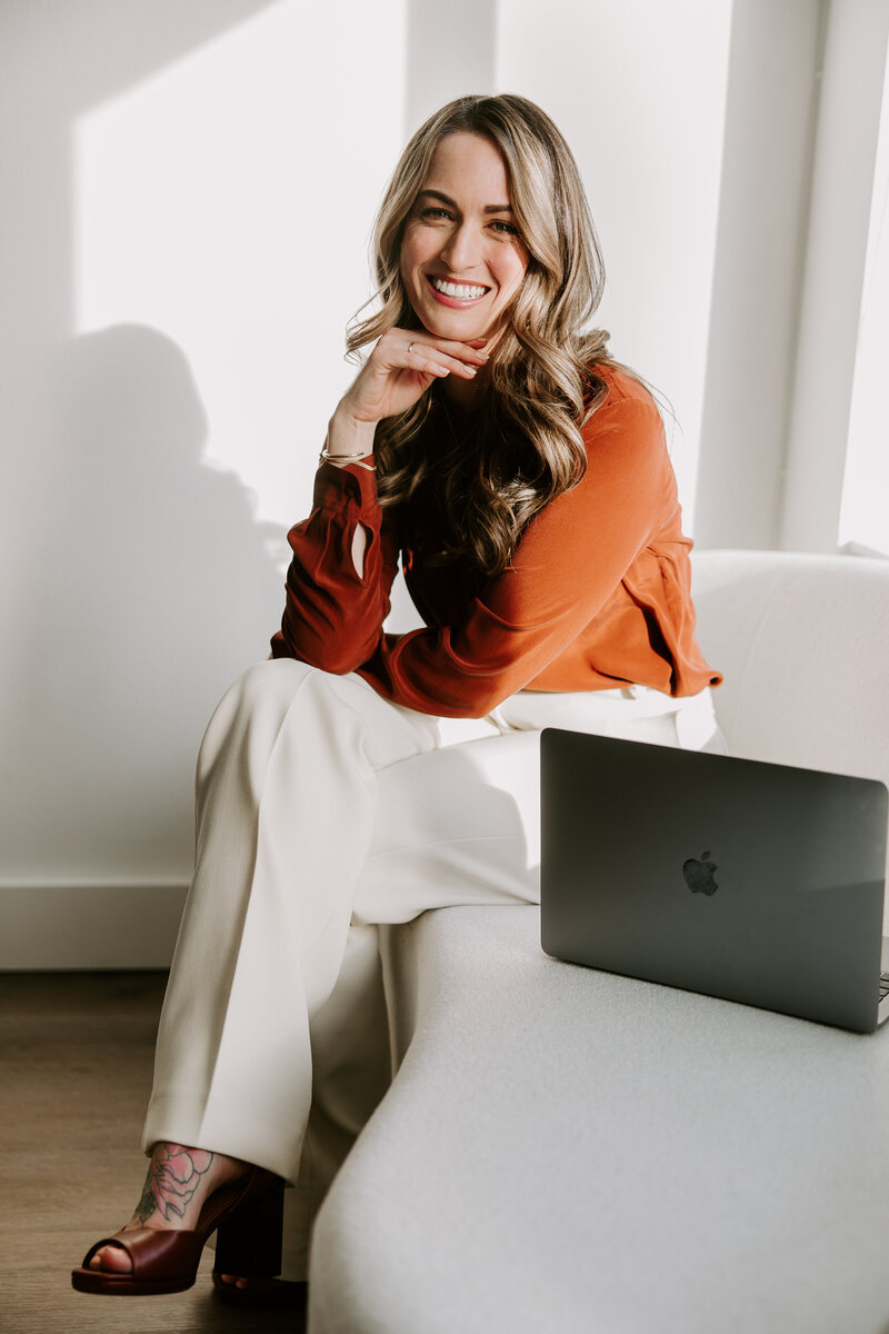 A studio portrait of founder and lead website copywriter Erika Fitzgerald, sitting on a white couch next to a laptop 