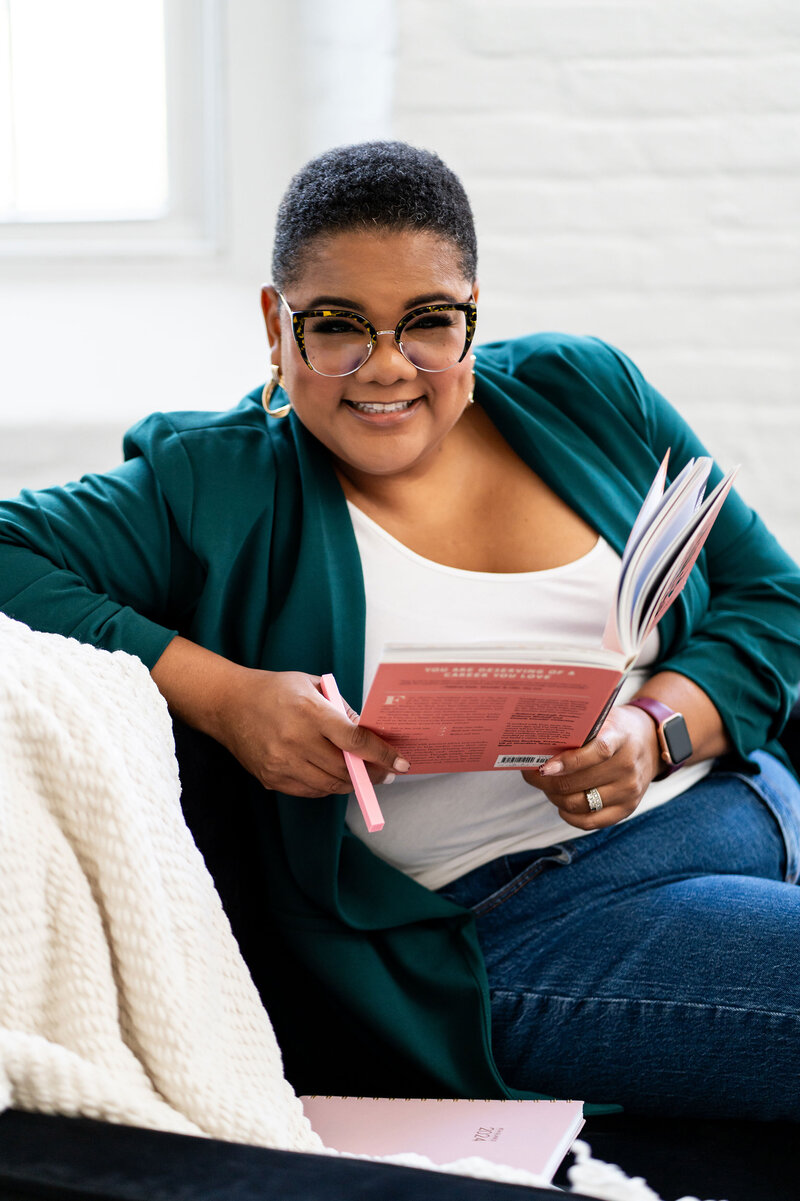 Group coaching for women's career development, featuring a professional wearing a branded color blouse and reading glasses, holding an important leadership book, in her office setting. This image represents empowerment, growth, and leadership for women in the workplace.