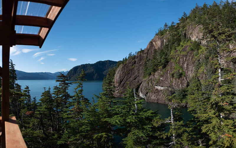 Looking out from the top of a lush green ridge in Kenai Fjords, the wooden edge of a luxury glamping yurt is visible in the edge of the frame with sweeping views of bright blue sky and rich blue water of Resurrection Bay during a luxury elopement in Kenai Fjords.
