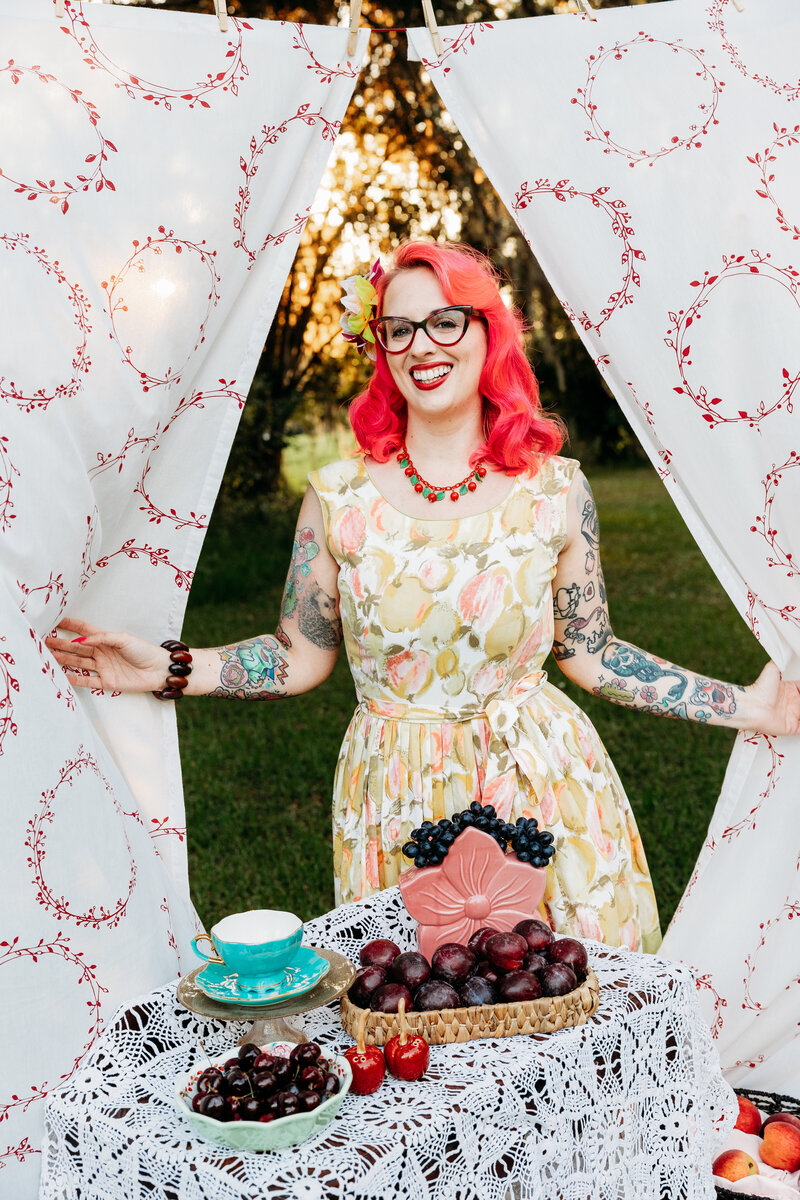 Woman poses playfully behind cherry-themed curtains at sunset, dressed in a retro yellow floral dress with bold pink hair.