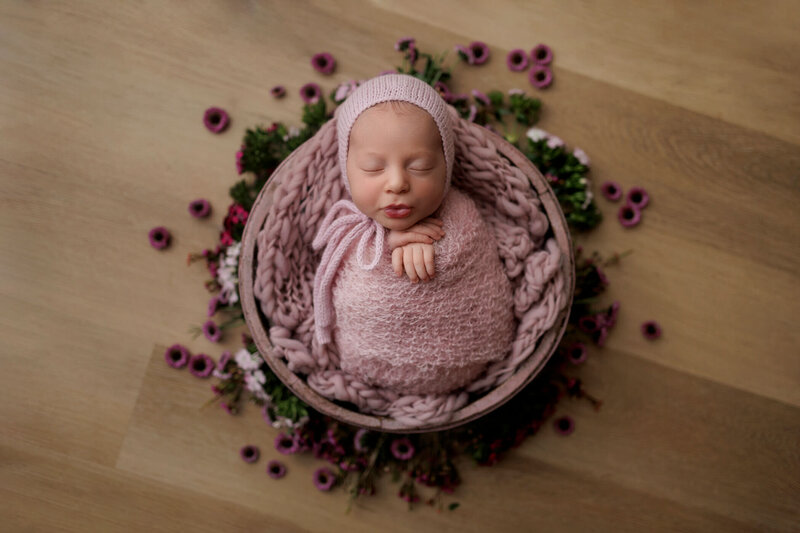 A newborn baby is peacefully sleeping, swaddled in a soft, pink knit blanket and wearing a matching pink bonnet. The baby is nestled in a cozy basket lined with a chunky knit blanket of a similar pink hue. The basket is placed on a wooden floor and surrounded by delicate purple flowers, creating a charming and serene setting. The baby's hands are gently crossed over their chest, and their expression is calm and content, making this a heartwarming and beautifully composed portrait.