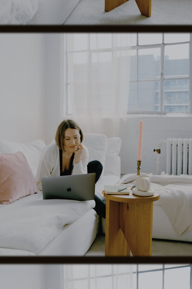Margarita. a website copywriter for creatives, sits on a white couch in a white cardian, looking at her laptop as if she is talking to someone on video. A white curtain gracefully hands over tall windows in the backdrop. Snow is gathered on the window sill. All is well.