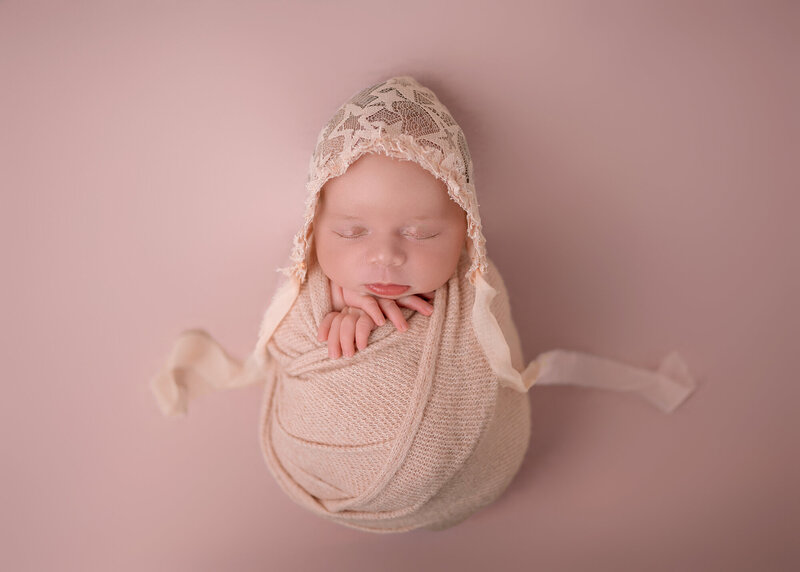 A newborn baby sleeps in a lace bonnet on pink bed