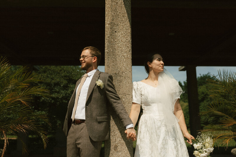 Bride and groom standing on either side of a column, holding hands before their ceremony in natural sunlight, captured by Rainstorm Photo & Video in an authentic documentary style.