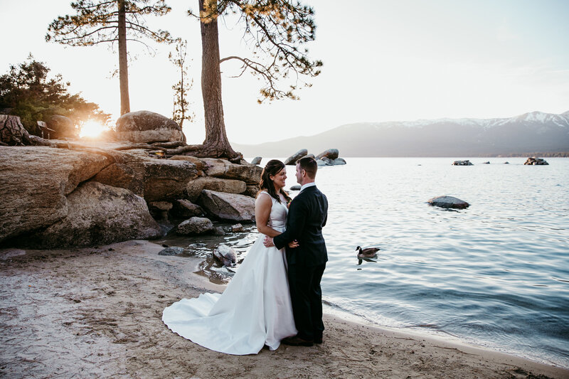 Bride and groom standing together at Sand Harbor Beach in Lake Tahoe during sunset, surrounded by rocks, pine trees, and calm water with mountain views in the background.